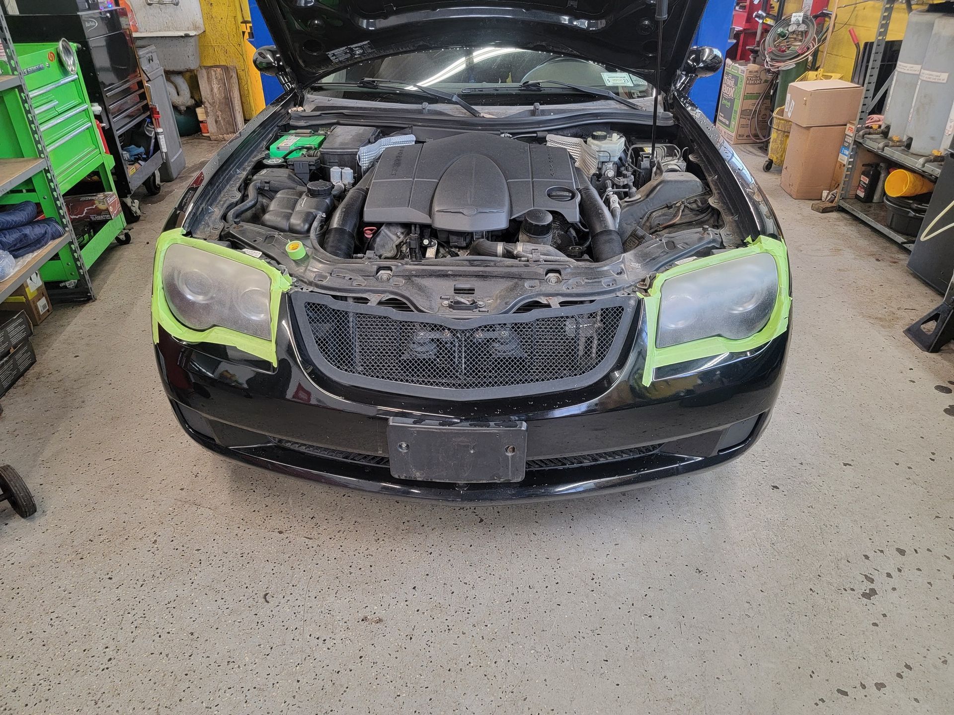 Black car with hood open, headlights taped off, in a garage.