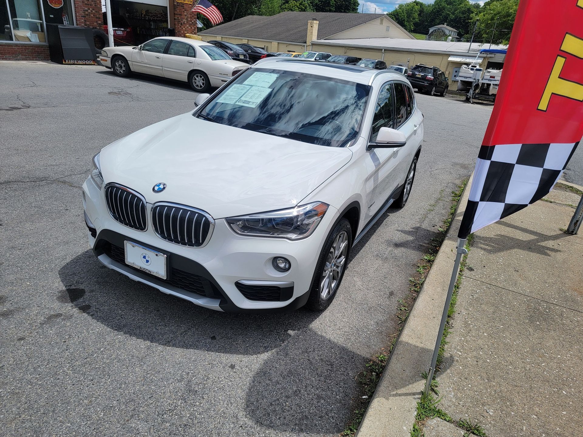 White BMW X1 SUV parked on asphalt, sunny day. Checkered flag and other cars in background.