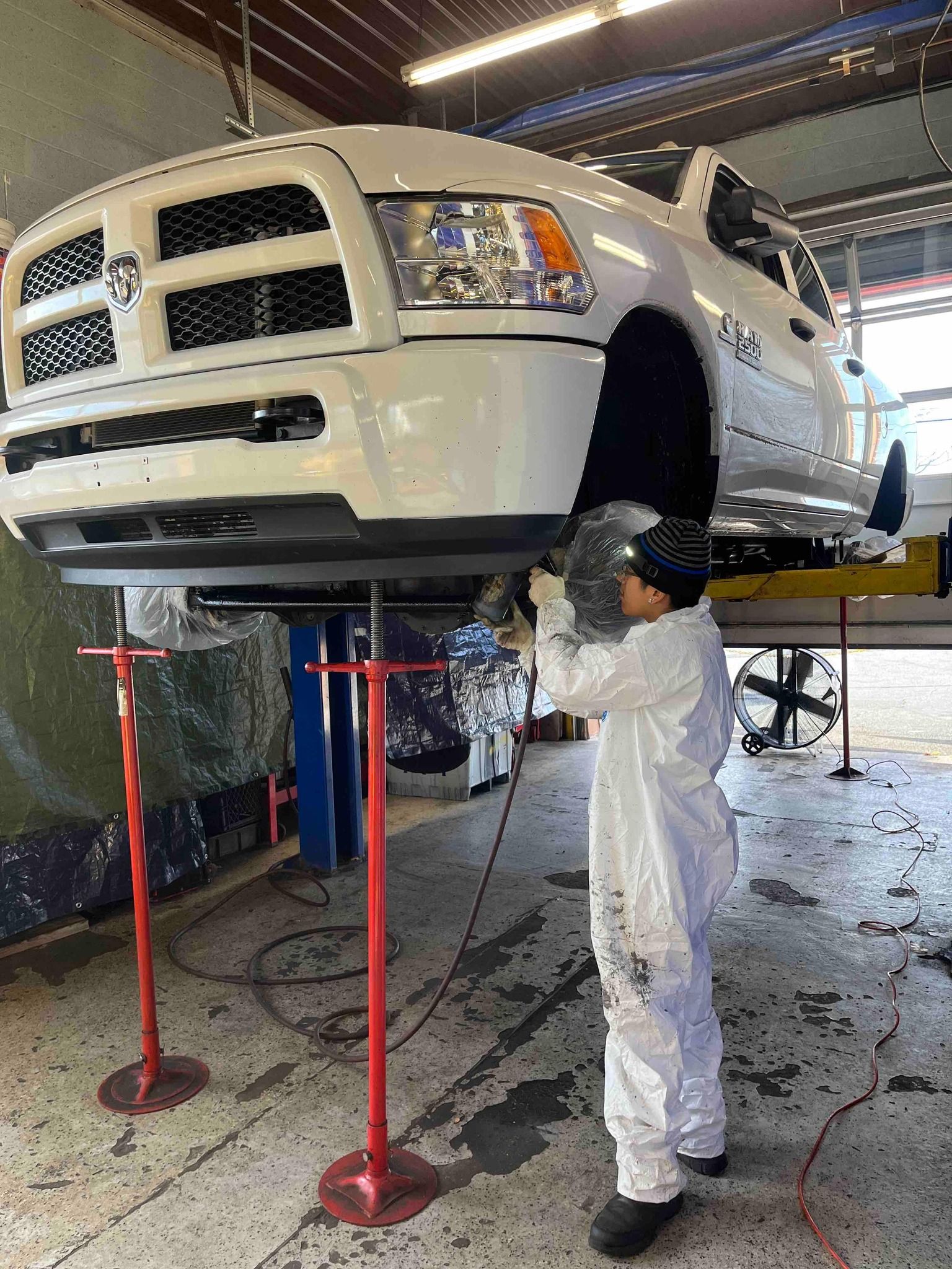 Mechanic in white coveralls works on a white Dodge Ram pickup truck raised on a lift in a shop.