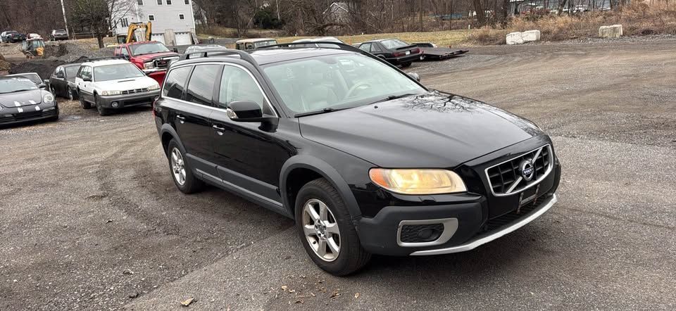 A black Volvo XC70 station wagon parked on a gravel lot with other vehicles visible in the background.
