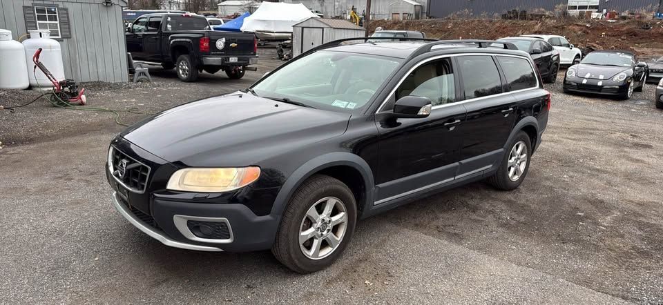 A black Volvo XC70 station wagon parked on a gravel lot near other vehicles.
