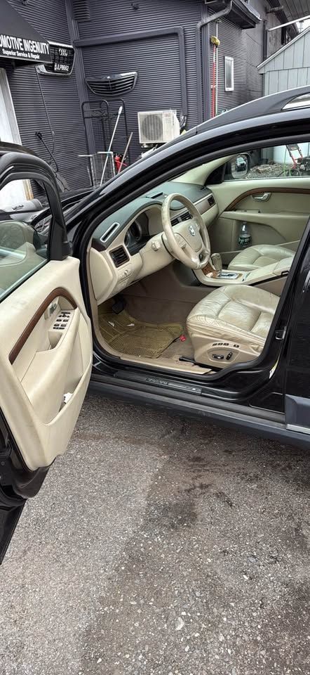 An open driver-side door reveals a black Volvo SUV with a light-tan leather interior, parked on a paved surface.