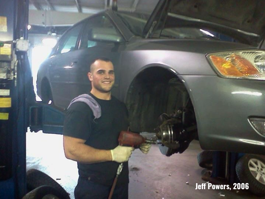 Mechanic smiles while working on a car in a shop. The car is gray and raised on a lift.