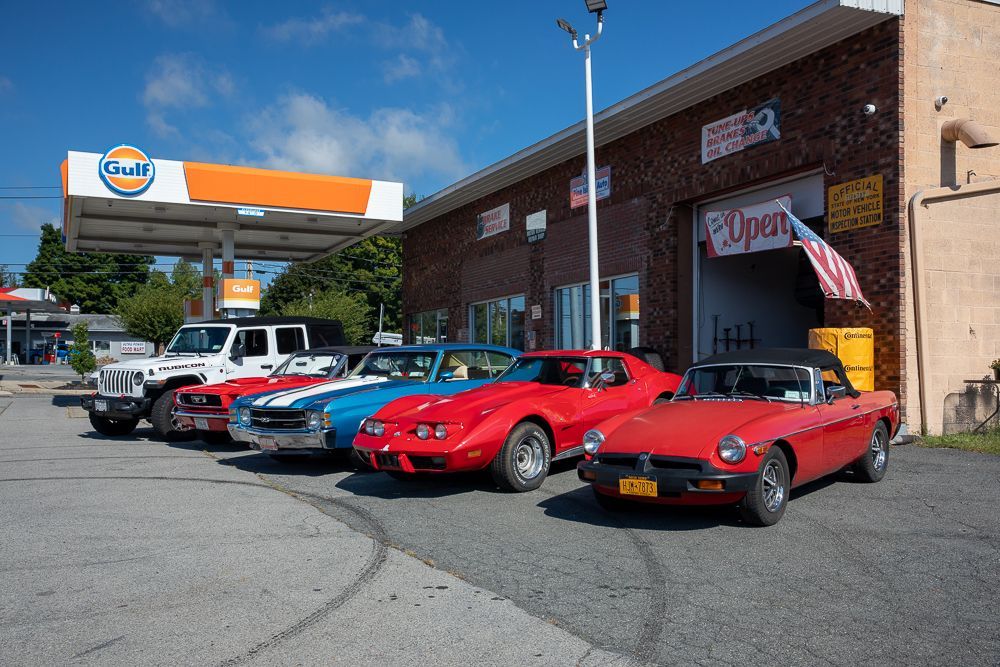 Classic cars parked in front of a brick building and a gas station on a sunny day.