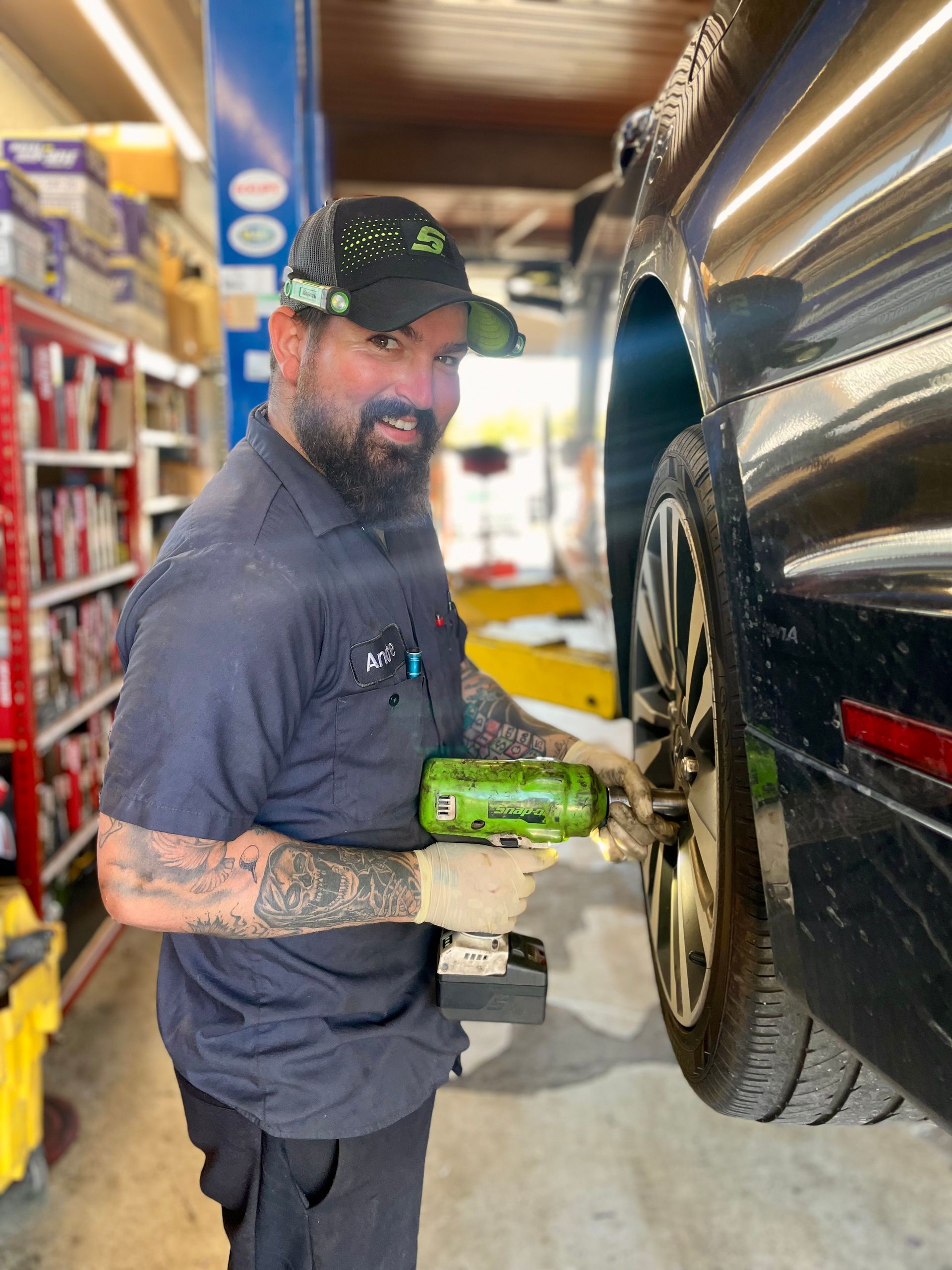 Mechanic uses a power tool on a car's wheel. He wears a hat, gloves, and a work shirt, smiles in a garage setting.