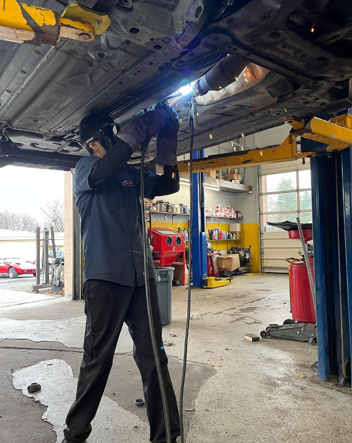 Mechanic welding under a car raised on a lift in a garage; sparks fly.