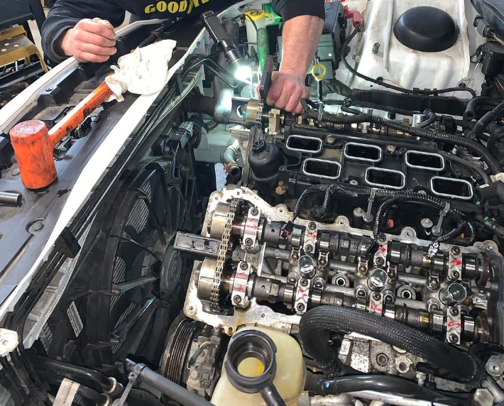 Two people working on a car engine with exposed components; setting is a garage.