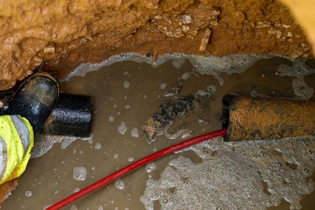 Flooded trench with muddy water, black pipe, and a red cable beside a worker’s boot