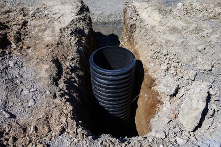 Black corrugated drainage pipe installed in a dug hole in rocky soil.