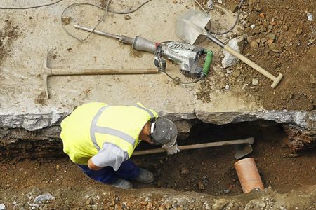Worker in a yellow safety vest inspecting a trench beside pavement with tools on the ground