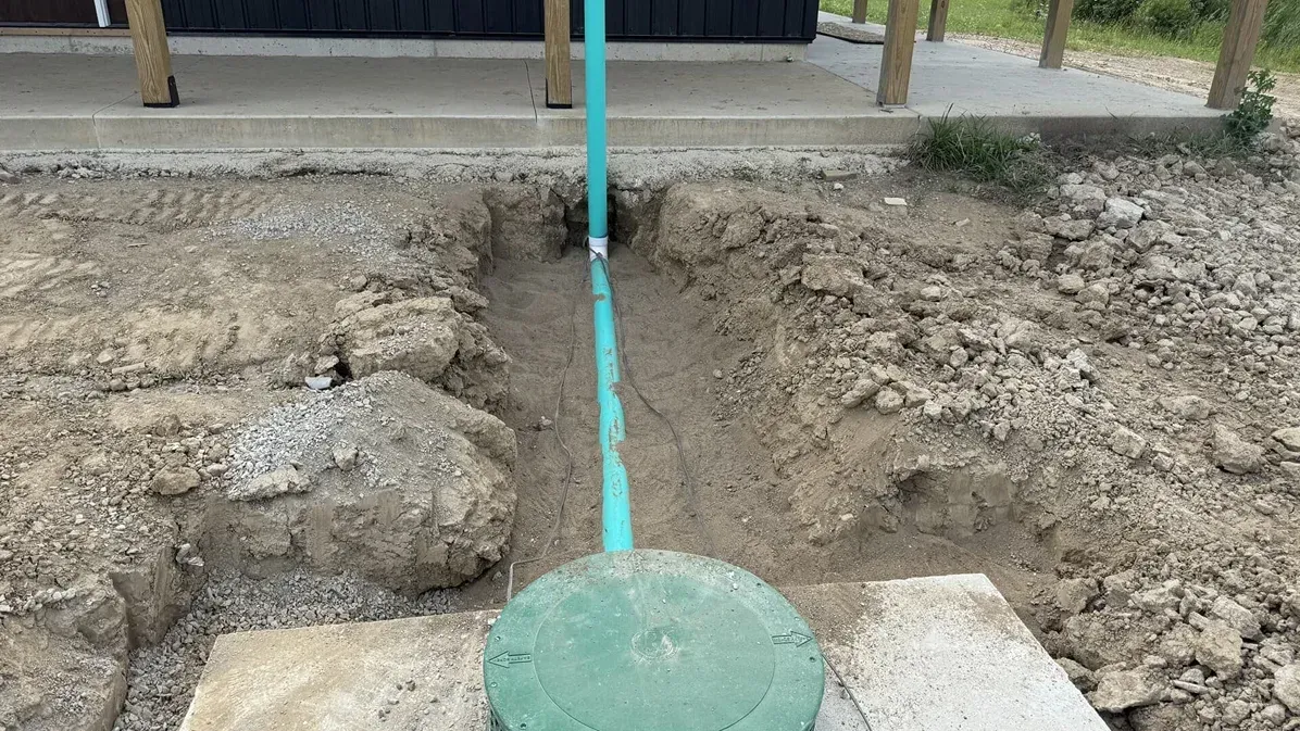 Excavated dirt area with a turquoise utility pipe running to a green circular cover near a building.