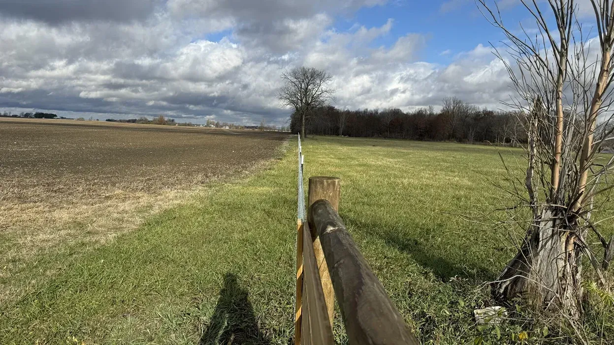 Rural field with a wooden fence, leafless trees, and cloudy sky over green and brown farmland.