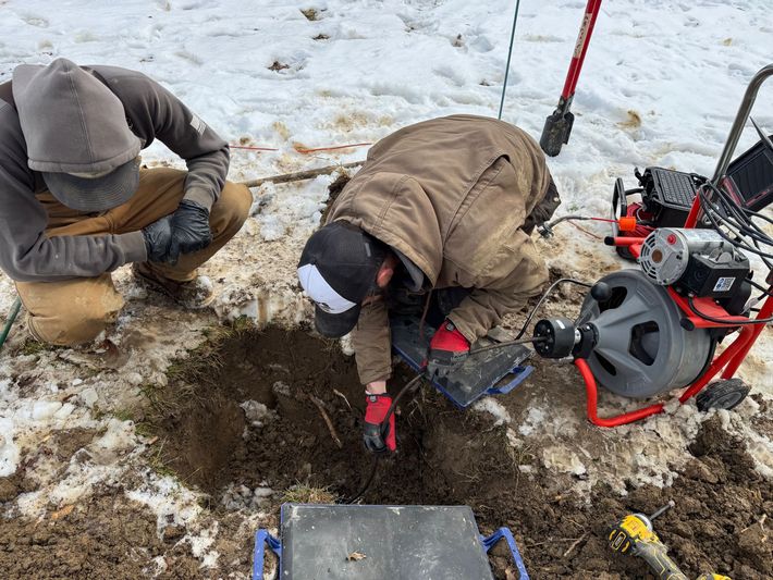 Two people kneeling in snow, digging a hole beside drilling equipment and tools.