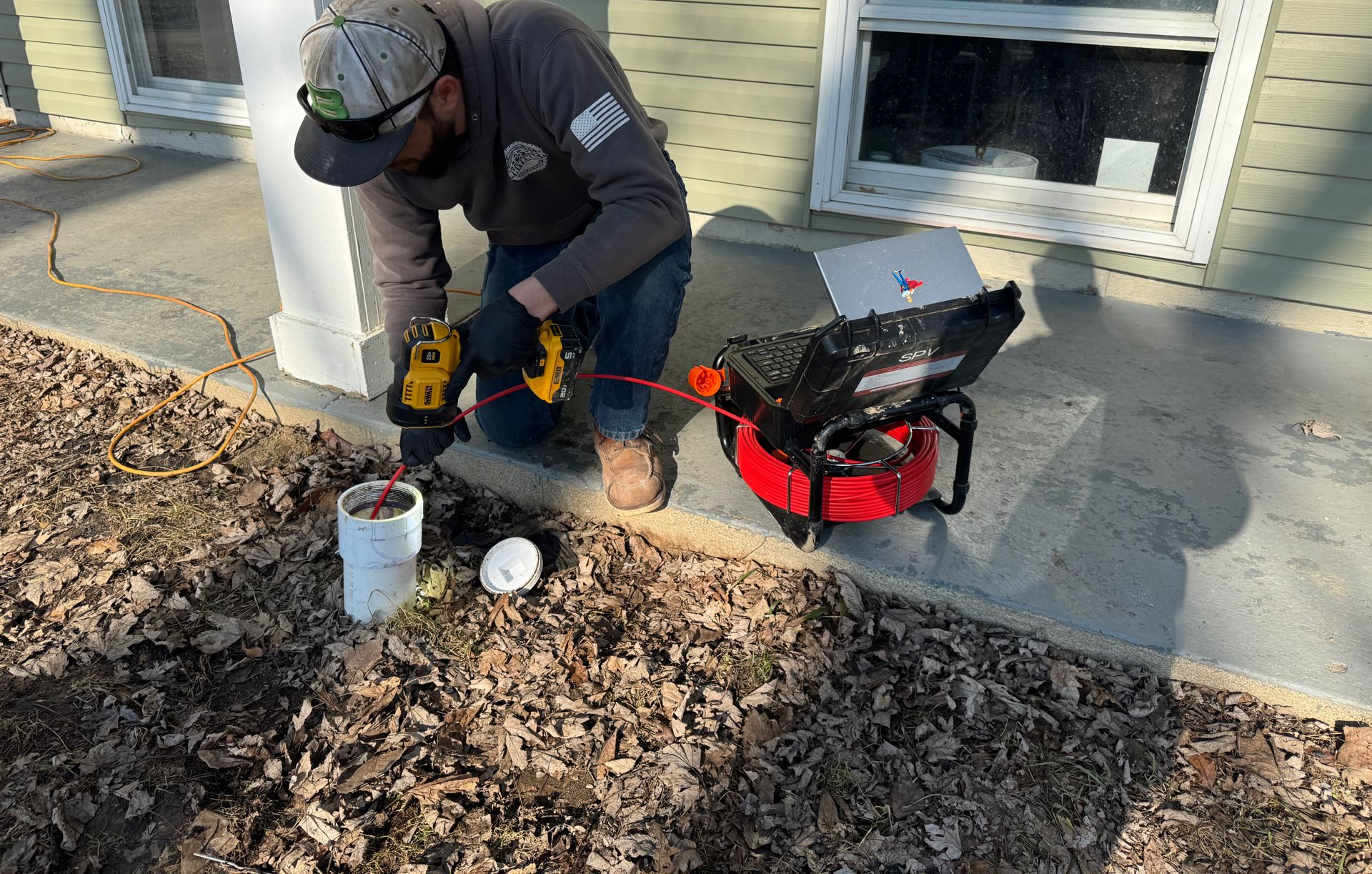 Worker using a drill to clean a gutter beside a house, with leaves piled on the ground.