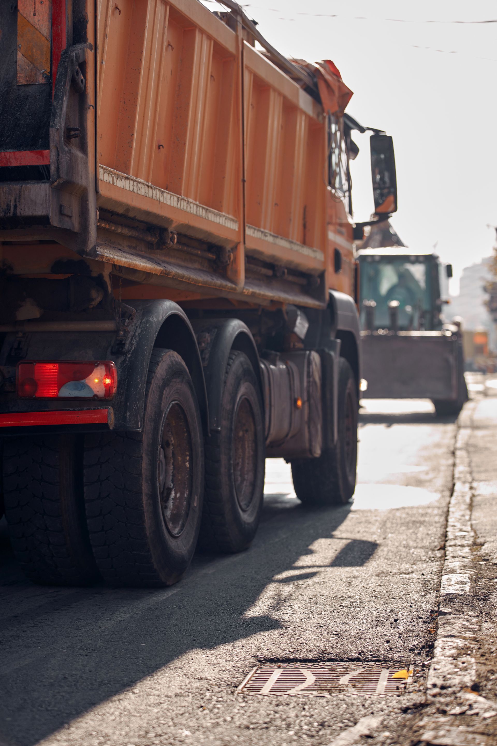 Un camion-benne orange est stationné sur une route goudronnée, une excavatrice étant visible en arrière-plan.