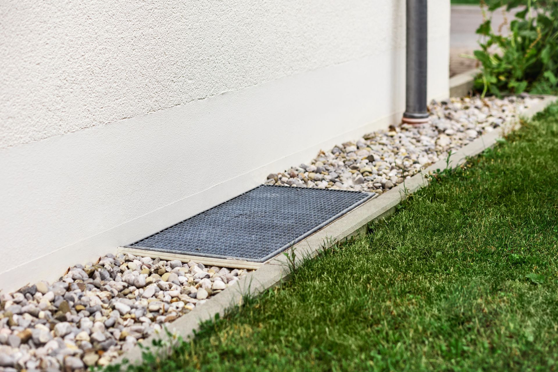 Une étroite grille métallique est posée sur un lit de gravier, contre le mur blanc d'une maison, près d'un tuyau de drainage et d'une pelouse.