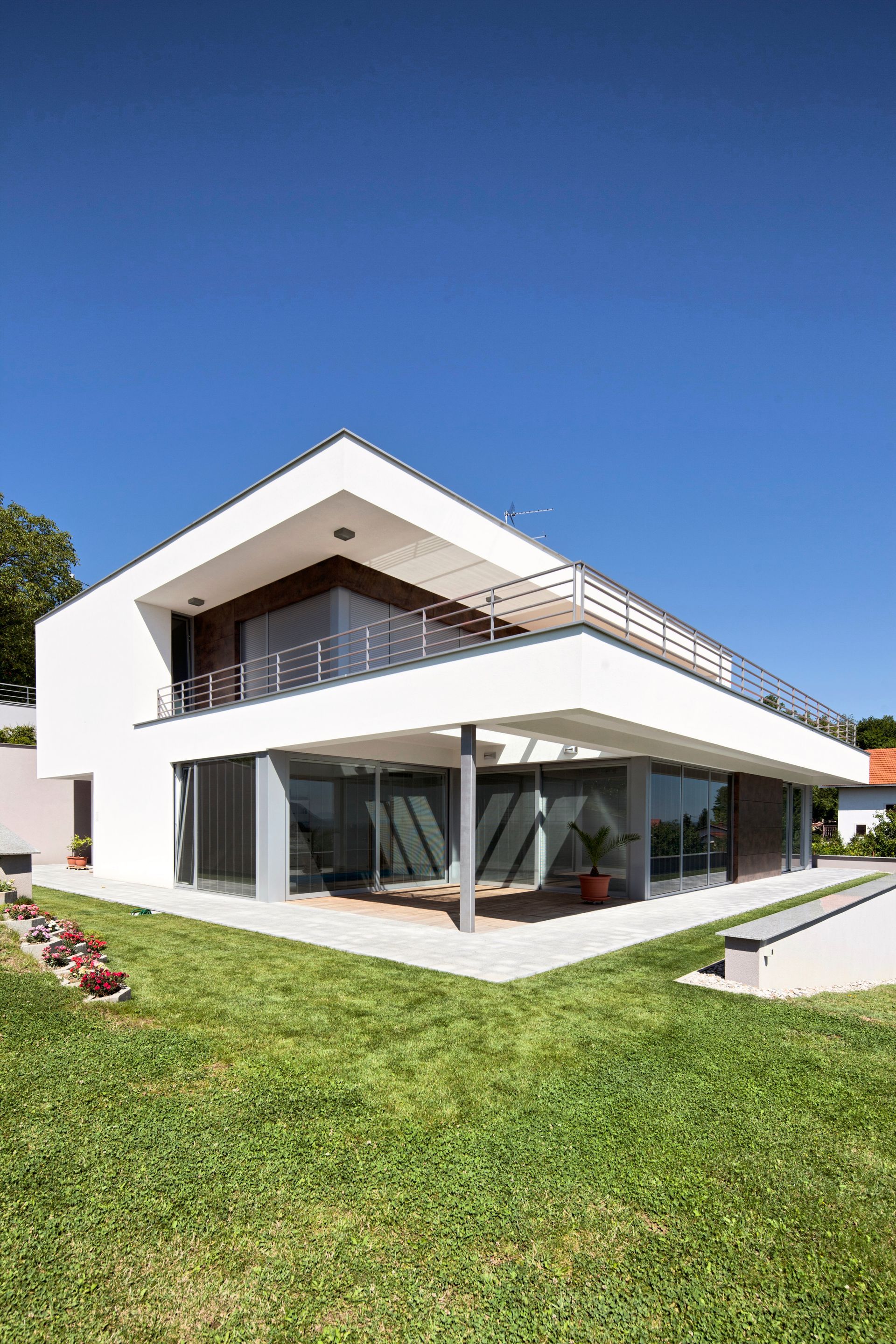 Une maison moderne, blanche, de deux étages, avec un toit plat, de grandes baies vitrées et un balcon donnant sur un ciel bleu azur.