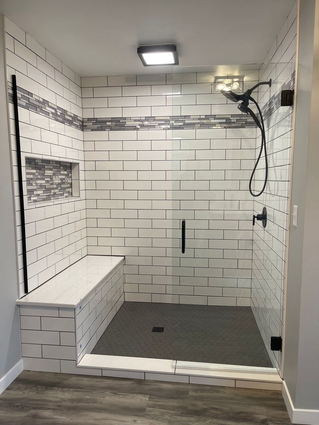 Tiled shower stall with bench, overhead light, dark floor, and wall-mounted shower fixtures