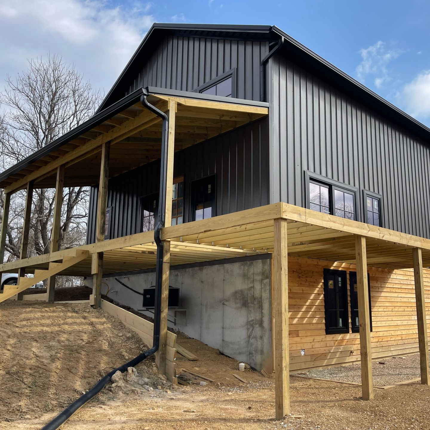 Black metal house on a sloped lot with unfinished wooden decks and a concrete foundation