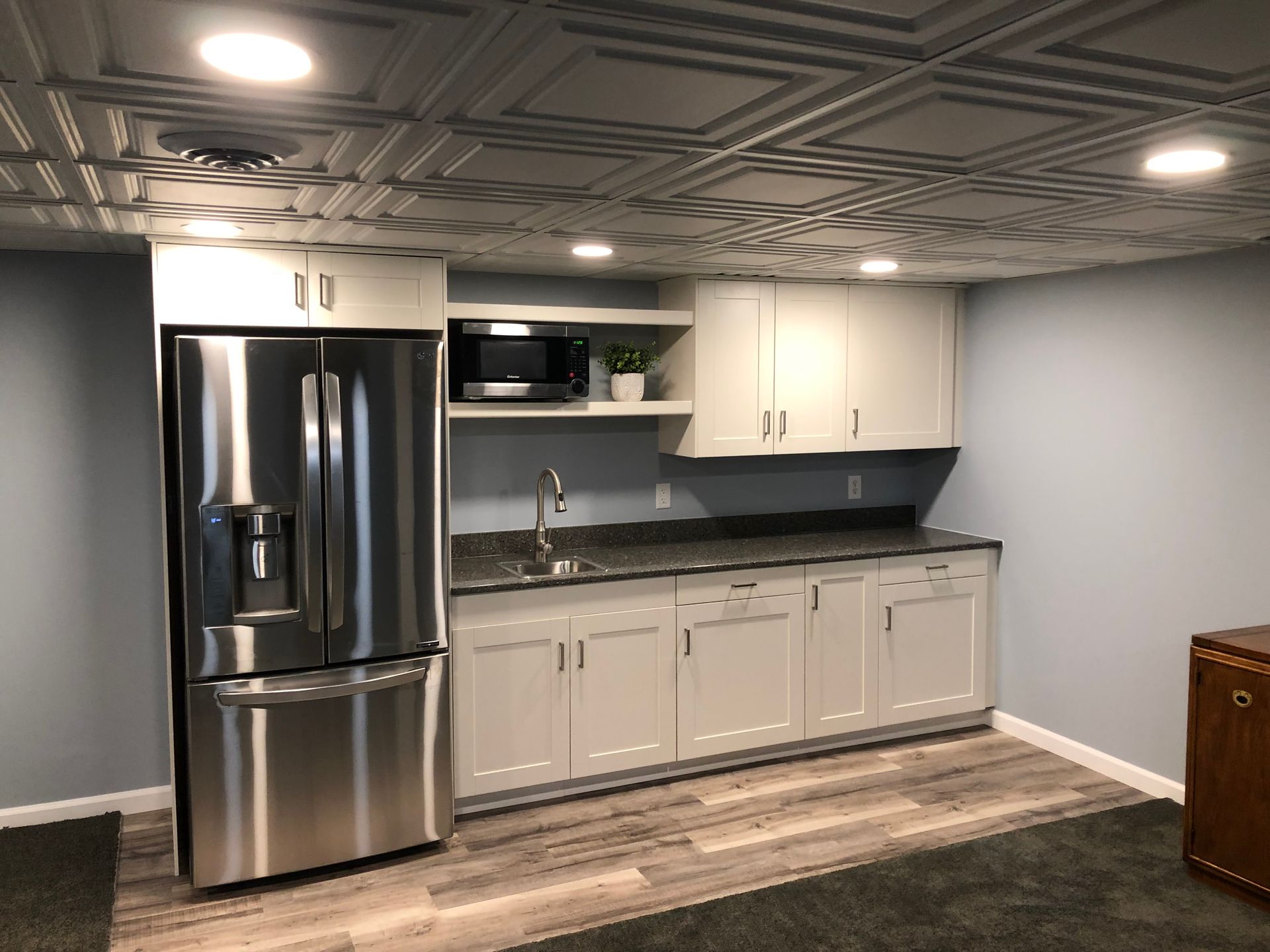 Modern kitchenette with stainless steel refrigerator, white cabinets, black countertops, and gray walls.