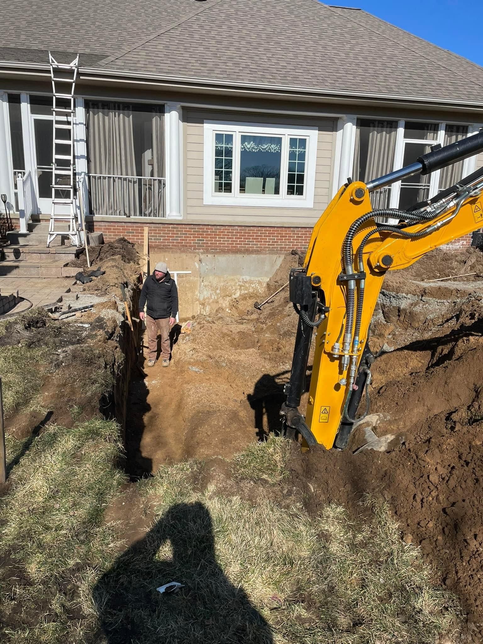 Person standing in a deep trench beside a yellow excavator in front of a house