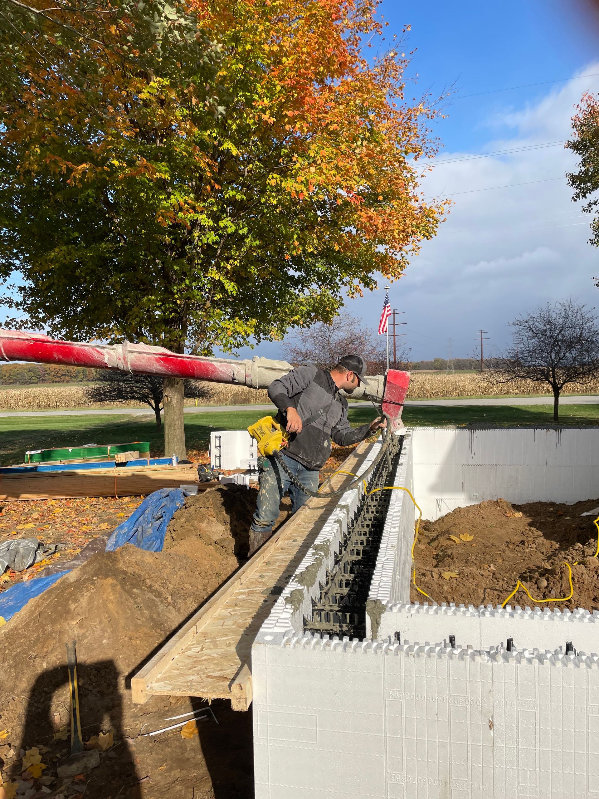 Two workers building a cinder block wall beside a red concrete pump under a tree on a sunny day