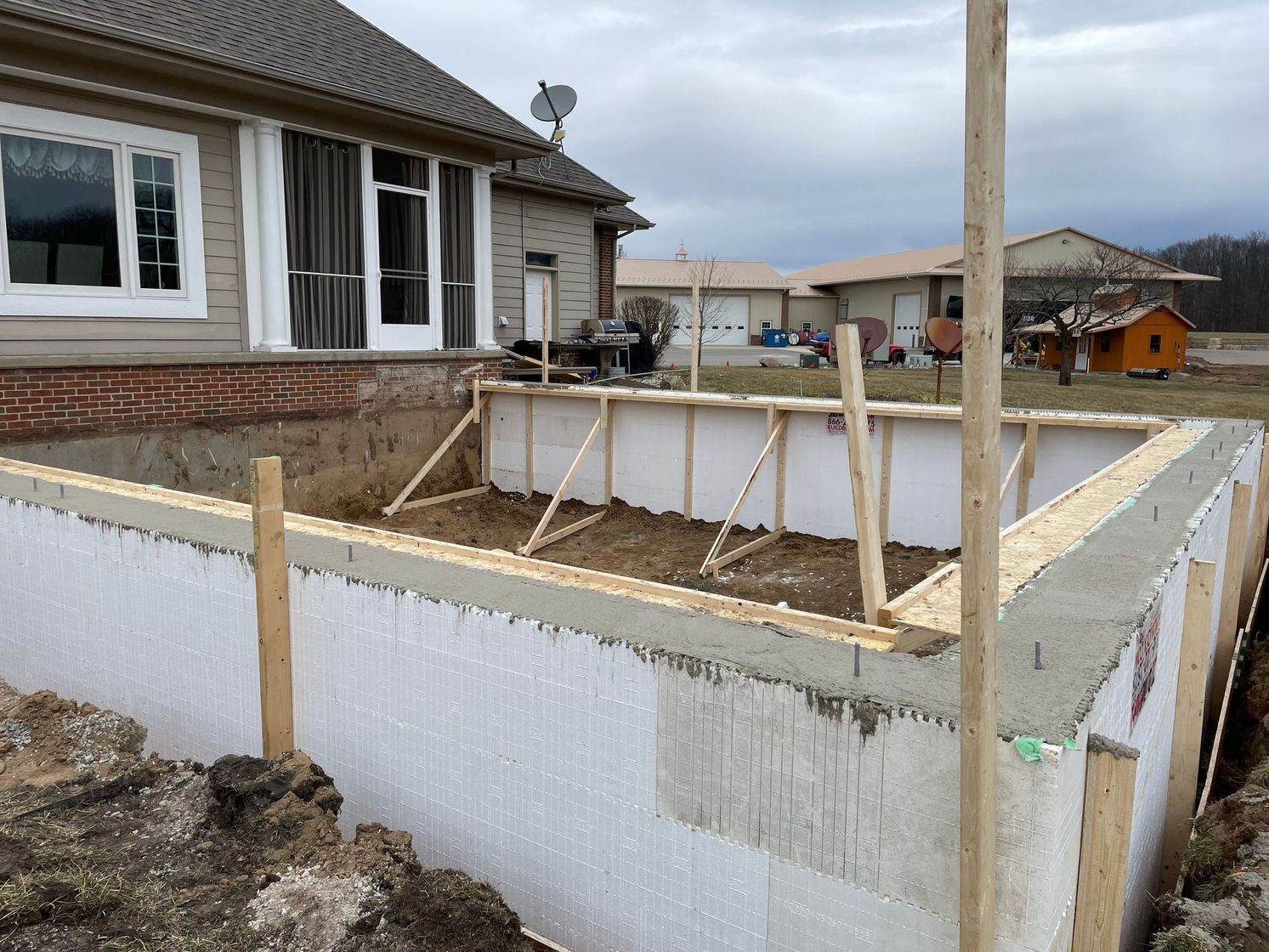 Partially built concrete foundation beside a house, with wooden supports and soil around the edges.
