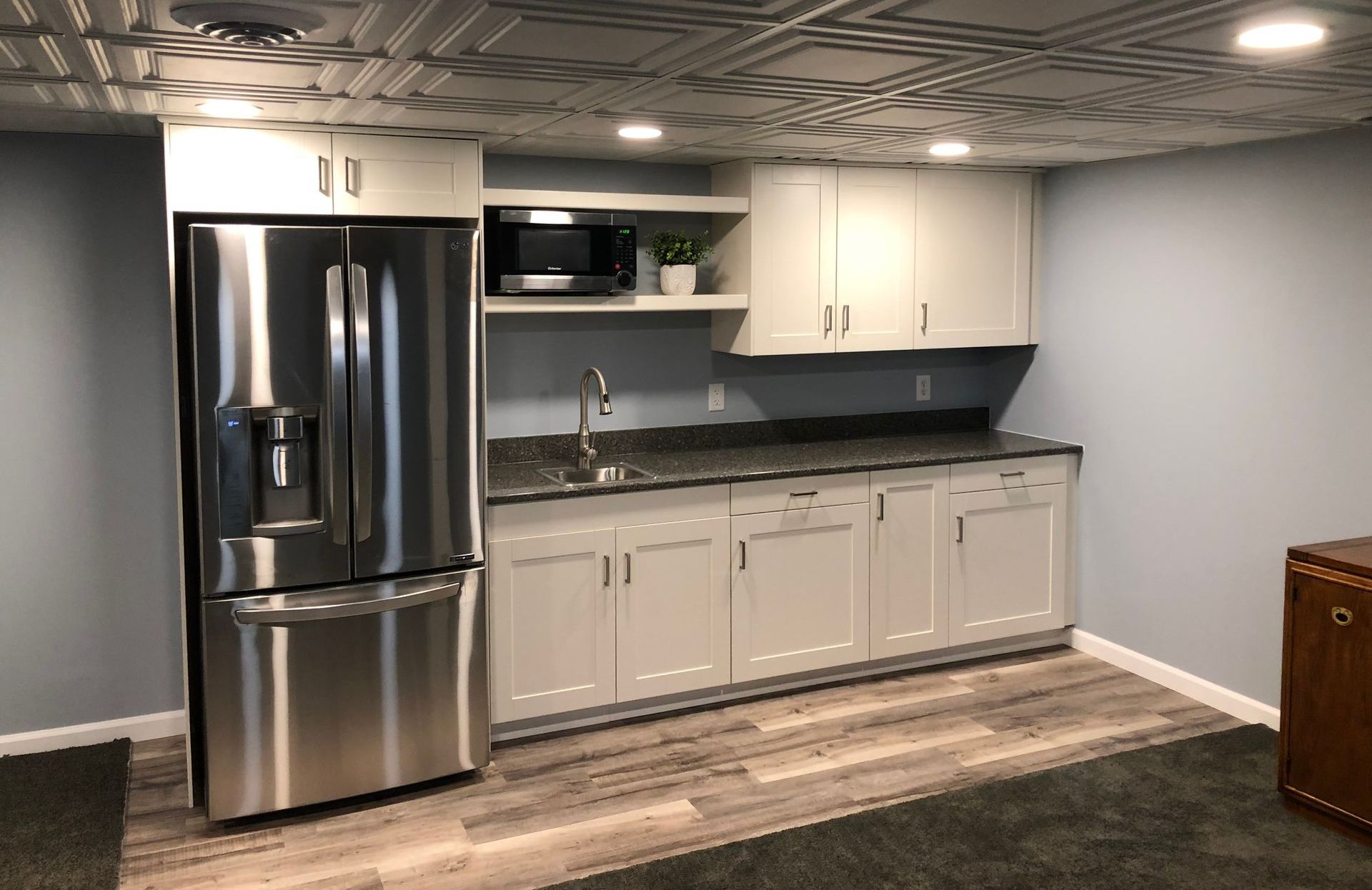 Modern basement kitchenette with stainless steel fridge, white cabinets, sink, and dark countertop
