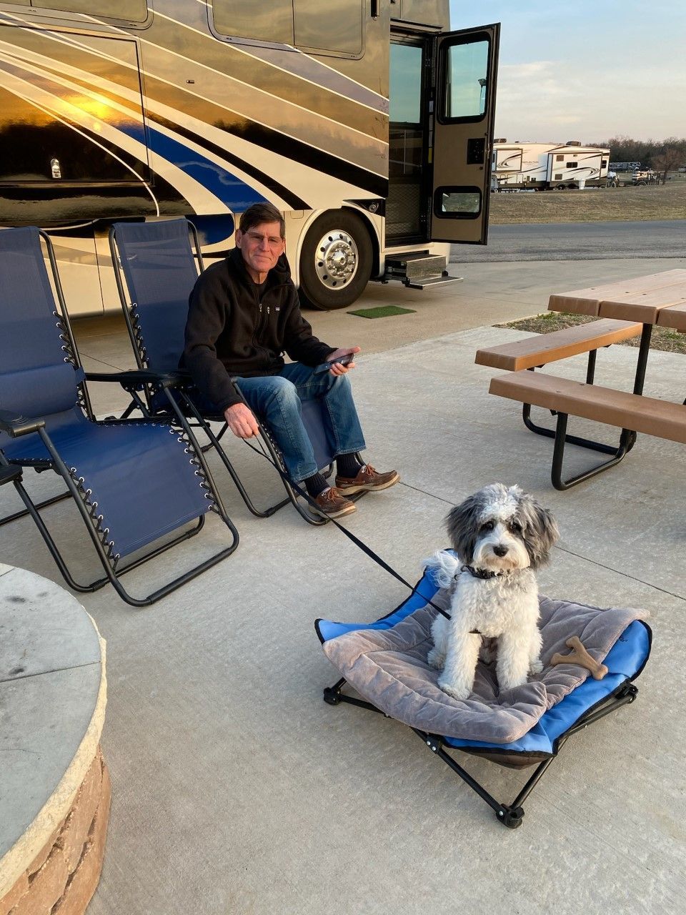 A man is sitting in a chair next to a dog on a bed.