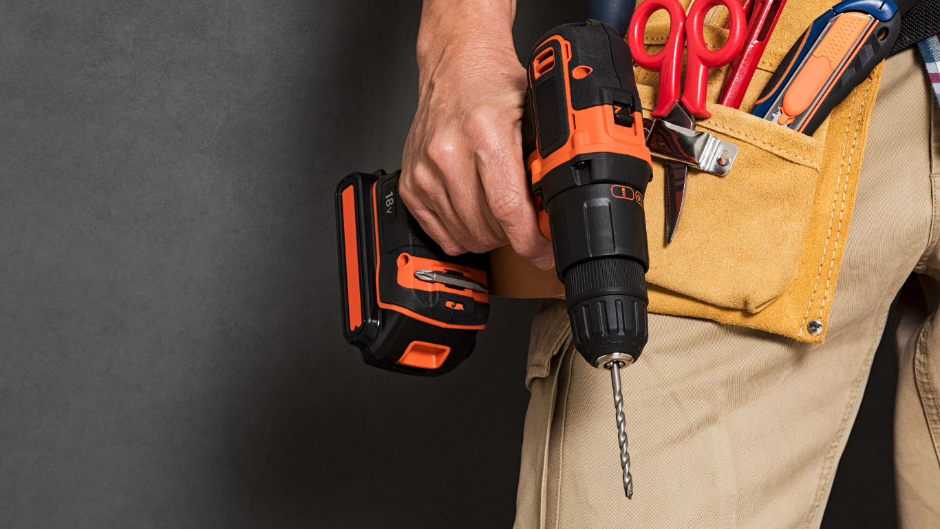 Person holding a cordless drill and tool belt with pliers and scissors, against a gray background