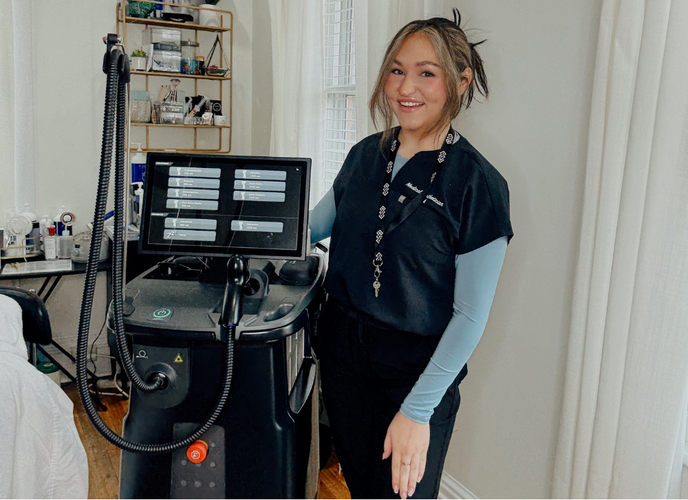 Woman in black uniform smiles next to a laser hair removal machine in a room with a window.