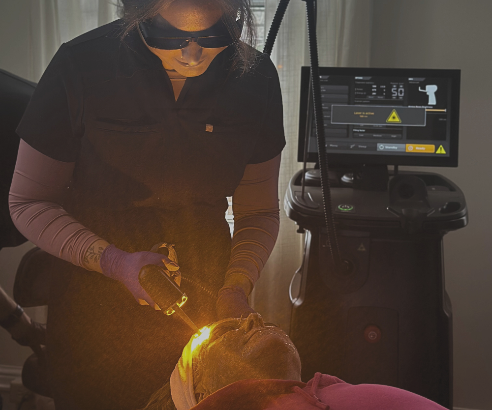 Woman in protective eyewear using laser device on a client's face in a medical setting.