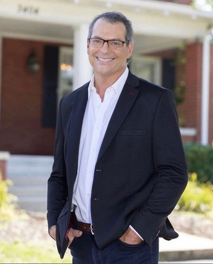 Man in a suit smiles, standing outside a brick house, hands in pockets.