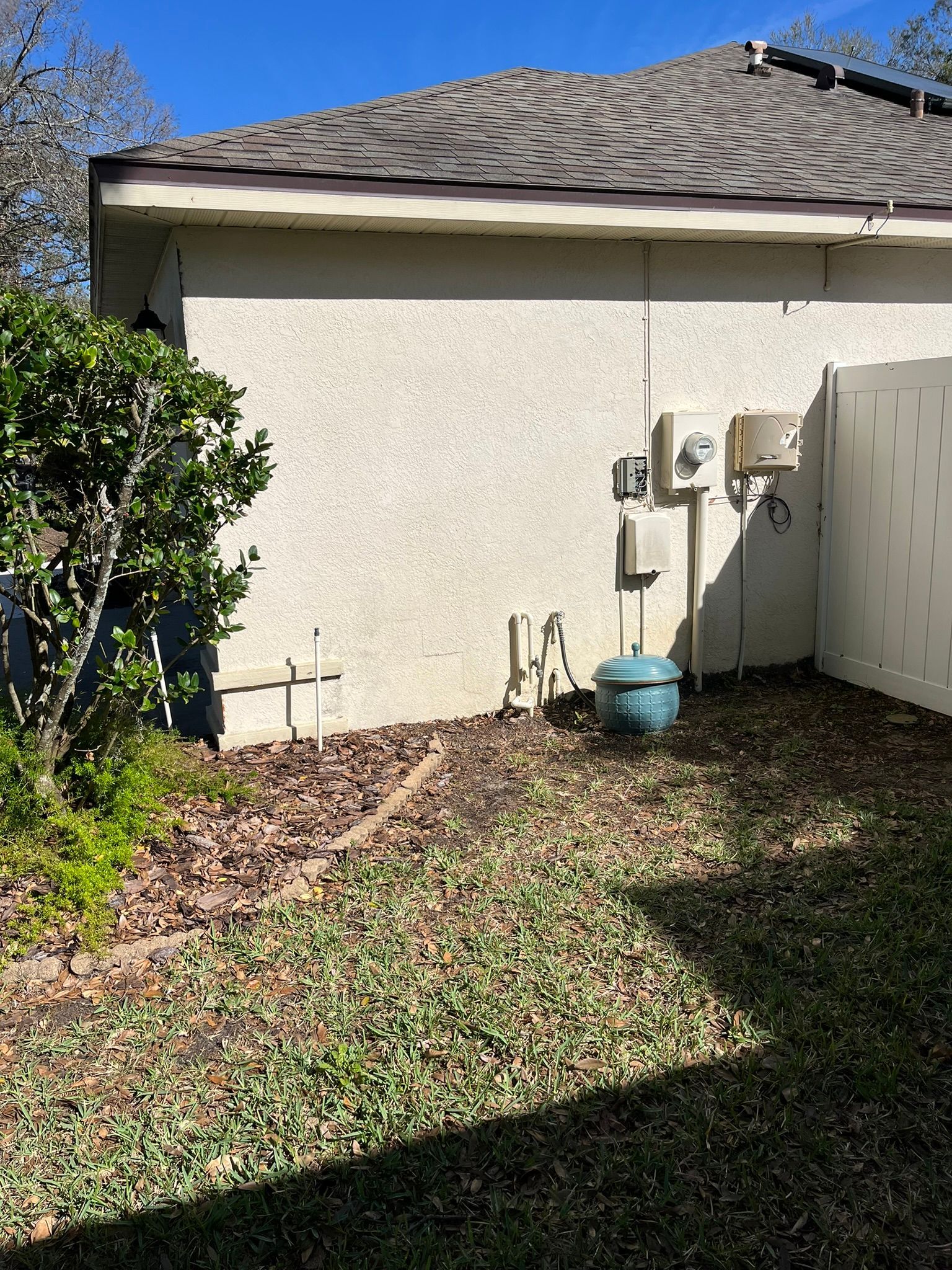 Backyard side of a house with utility meters, beige stucco wall, grass, and fallen leaves