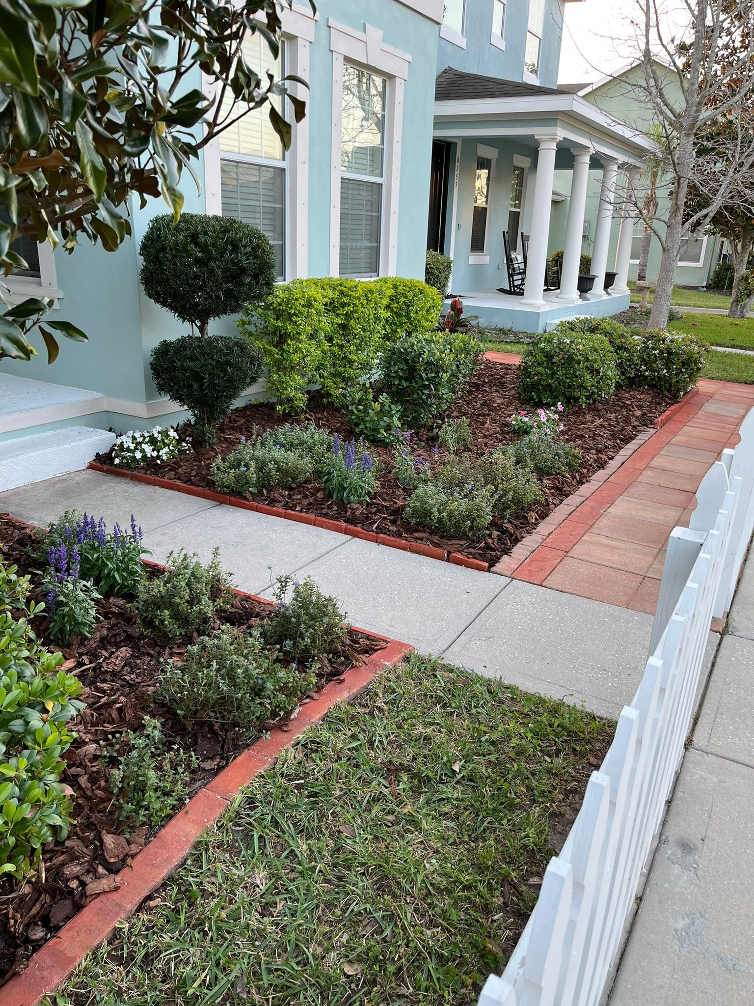 Front yard garden with trimmed shrubs, brick edging, and a white house porch along a sidewalk