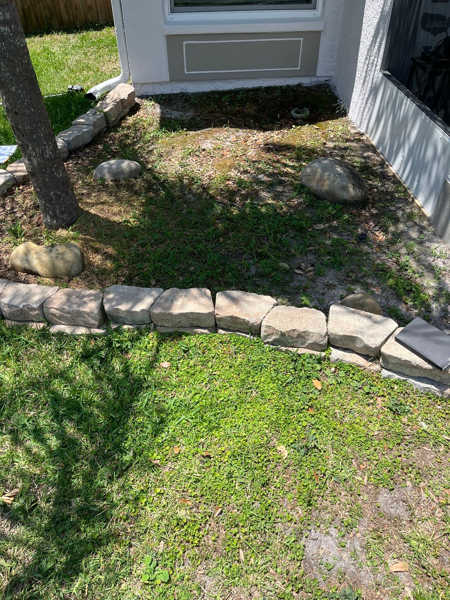 Small front yard garden bed with rocks, sparse plants, and a stone border by a house entrance