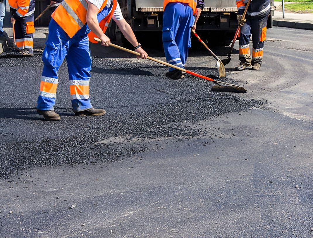 Road workers in blue and orange safety vests leveling asphalt with rakes on a paved road.