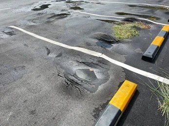 Damaged asphalt parking space with potholes, broken white lines, and yellow/black parking bumpers.