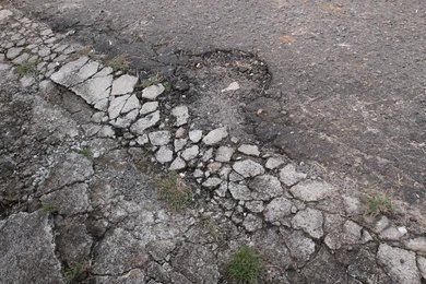 Cracked asphalt road with a line of broken concrete, pothole in the background, gray tones.