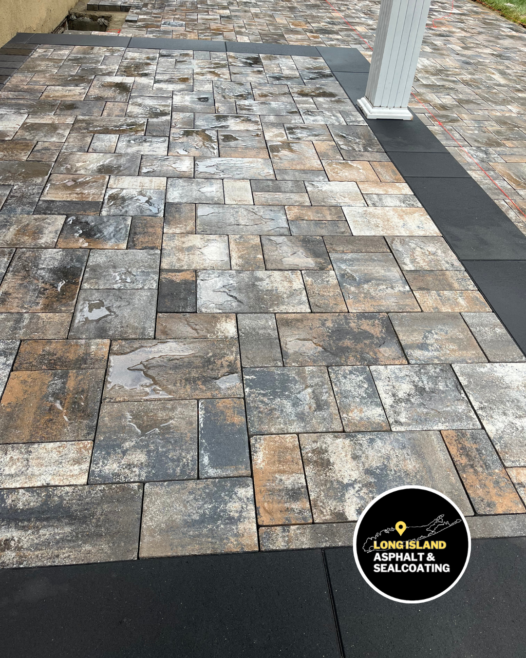 Patio paved with gray, brown, and black interlocking pavers. Black border runs around edge.