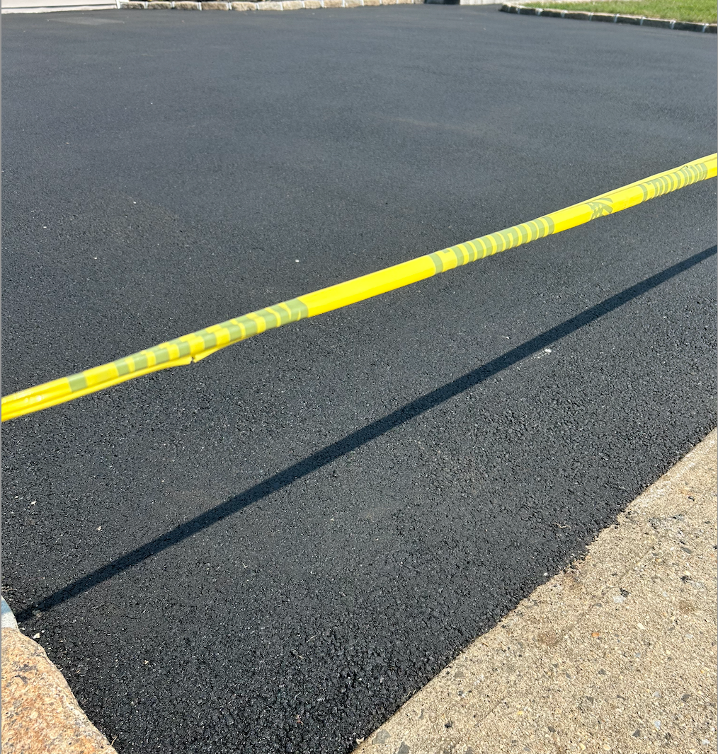 Freshly paved black asphalt with yellow caution tape, next to a grassy area.