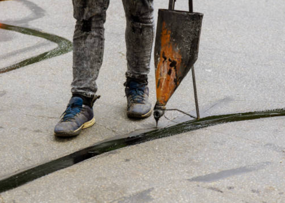 Person filling a crack in concrete with a sealant applicator; black sealant, grey jeans, asphalt setting.
