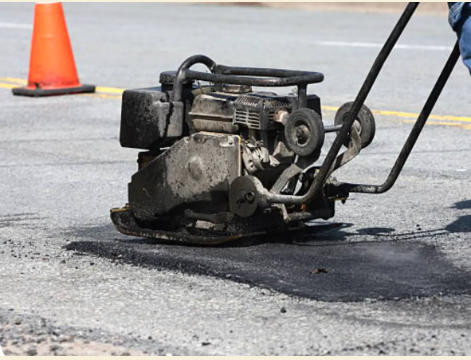 Person operating a plate compactor on asphalt road, compacting dark patch near a traffic cone.