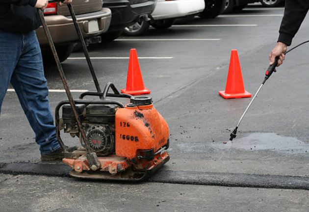 Two people repairing asphalt in a parking lot, one using a compactor and the other spraying material.