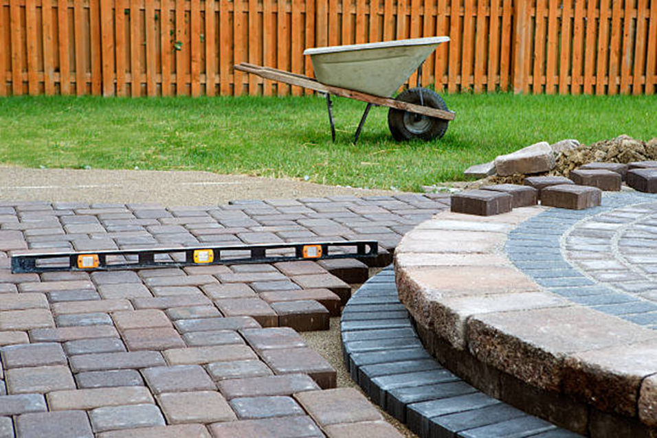 Brick patio construction in a backyard, with a level and wheelbarrow visible.