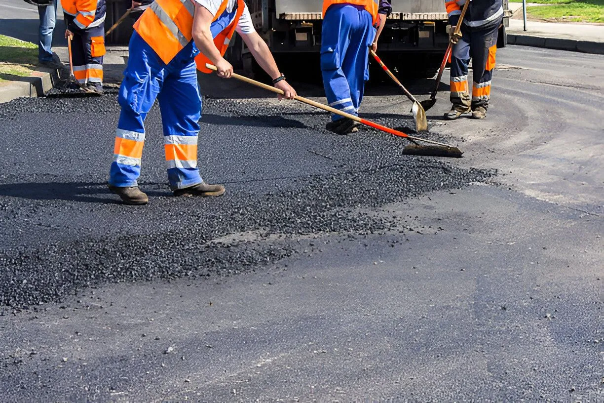 Road workers in blue coveralls and orange vests paving asphalt.