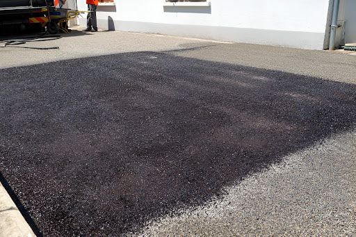 A freshly paved square of asphalt on a concrete surface, with a worker standing nearby.