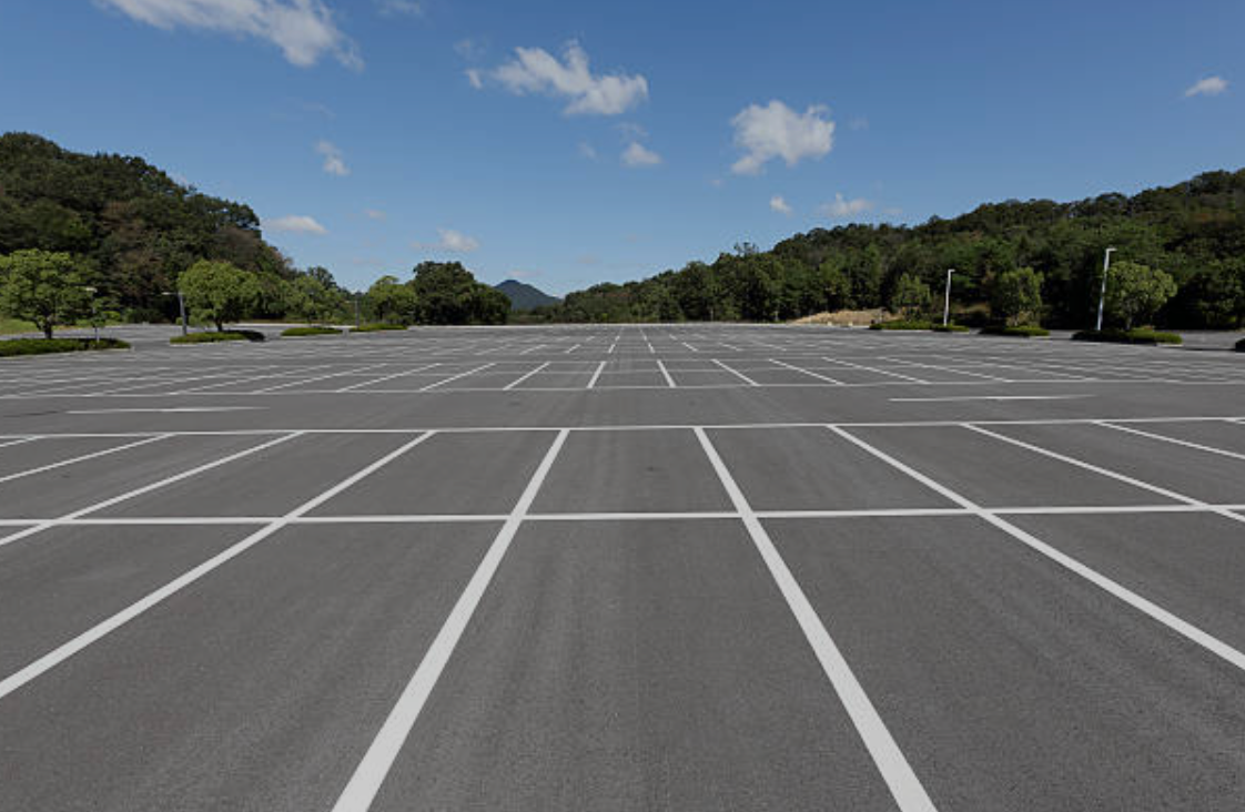 Empty asphalt parking lot with white painted lines under a blue sky, trees in the background.