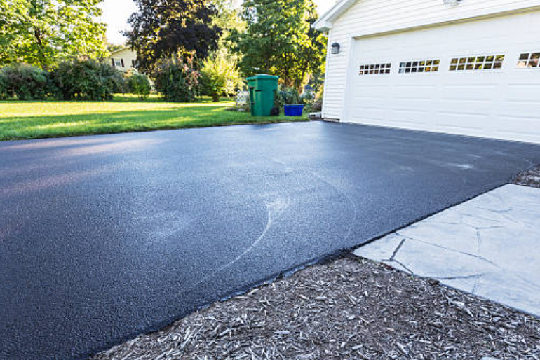 Newly sealed black asphalt driveway next to a white garage, green lawn, and mulch.
