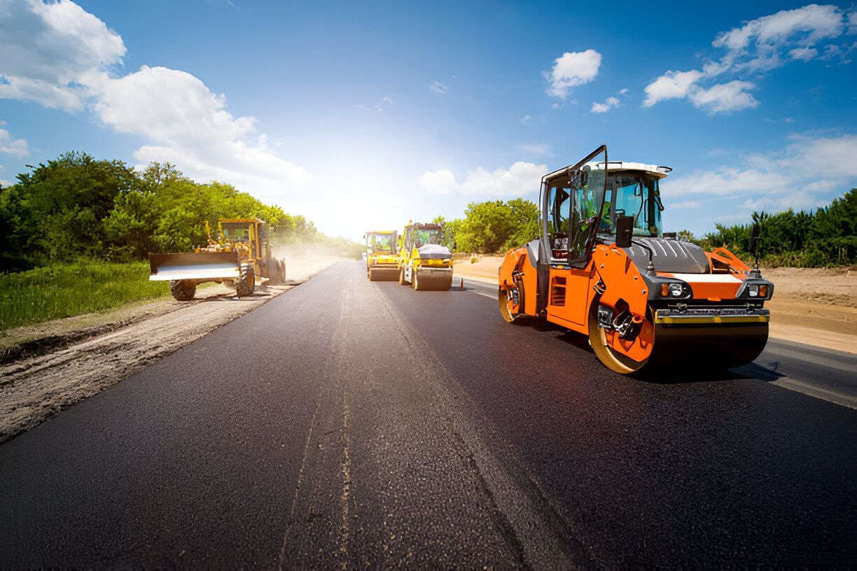 Road construction: Asphalt being laid with heavy machinery including a roller and bulldozer on a sunny day.